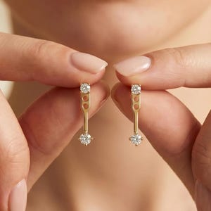 May include: A pair of gold drop earrings, each featuring a vertical bar design with a small, clear, round-cut gemstone set at the top and a matching, slightly larger, clear, round-cut gemstone dangling at the bottom of the slender gold post.