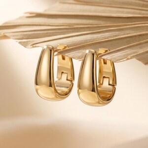 May include: A pair of polished gold-tone hoop earrings with a smooth, rounded, teardrop-shaped profile. The earrings feature a hinged closure mechanism, showcasing a reflective metallic finish and a clean, solid construction throughout the design.