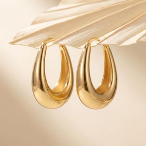 May include: A pair of polished gold hoop earrings with a smooth, teardrop-shaped silhouette. The earrings feature a thick, rounded metal construction with a high-shine finish, designed as open hoops with a secure post closure at the top.