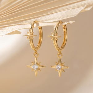 May include: A pair of gold hoop earrings featuring a small fixed starburst charm on the hoop and a dangling starburst charm at the bottom. Each dangling charm is centered with a single clear round gemstone, creating a symmetrical design.