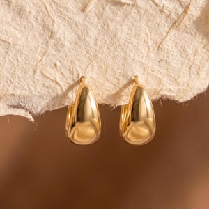 May include: A pair of small, polished gold hoop earrings with a smooth, teardrop-shaped profile. The earrings are suspended from a piece of textured, off-white handmade paper, showcasing a reflective metallic finish and a simple, curved design.