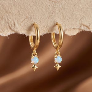 May include: A pair of gold hoop earrings featuring a small, round, iridescent white opal stone dangling from each hoop. Below each opal is a tiny, four-pointed star charm. The earrings have a polished gold finish and a secure hinged closure.