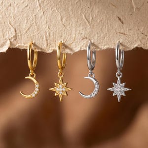 May include: Four hoop earrings featuring dangling charms. Two earrings are gold-toned, one with a crescent moon charm and one with a star charm. Two earrings are silver-toned, one with a crescent moon charm and one with a star charm. All charms are accented with small clear stones.