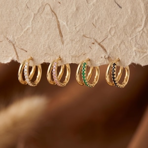 Könnte beinhalten: Vier goldene Creolen in einer Reihe angeordnet. Jeder Ohrring hat ein Doppel-Reifen-Design mit einem schlichten Goldband und einem mit kleinen Steinen besetzten Band. Die Steine sind jeweils klar, hellrosa, smaragdgrün und schwarz.