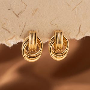 May include: A pair of gold-toned earrings featuring a design of three interlocking circular rings suspended from a vertically ribbed rectangular top piece. The earrings have a polished, reflective metallic finish and a structured, layered appearance.