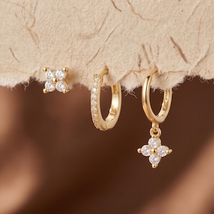 May include: A set of gold-toned jewelry consisting of one stud earring with a four-petaled flower design, one small hoop earring with a row of clear stones, and one hoop earring featuring a dangling four-petaled flower charm with clear stones.