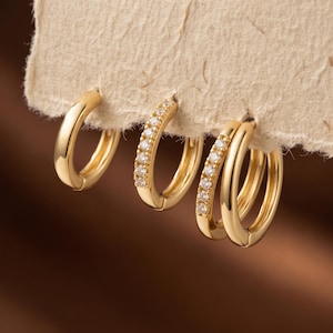 May include: A set of four gold hoop earrings displayed on a textured off-white paper card. One hoop is plain gold, one features a row of small clear stones, and two are linked together, with one plain gold and one featuring a row of clear stones.