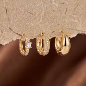 Peut inclure: Trois créoles dorées présentées en rangée. La première boucle d'oreille présente une pierre centrale carrée avec des pierres rondes plus petites sur l'anneau. La deuxième est pavée de petites pierres. La troisième est une créole dorée lisse et polie.