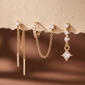 May include: Three gold-tone earrings featuring clear cubic zirconia stones. The first has two dangling chains with bar pendants. The second is a hoop connected to a stud by a chain. The third features three round stones and a square-cut drop.