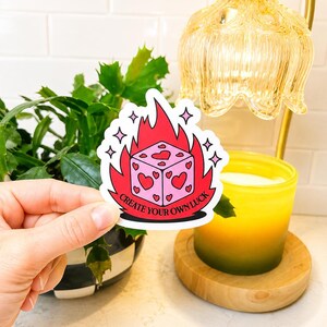 May include: A die-cut sticker featuring a light pink cube with red heart pips, surrounded by bright red flames and small pink star accents. The text "CREATE YOUR OWN LUCK" is printed in black capital letters along the bottom of the flame design.
