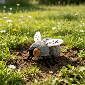 Può includere: Un peluche a forma di ape con un corpo grigio morbido e soffice, zampe e antenne nere. L'insetto ha occhi testurizzati color marrone chiaro e ali bianche con dettagli di venature cucite in marrone scuro, posizionato sul terreno marrone.