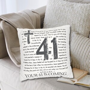 Könnte beinhalten: Ein quadratisches weißes Dekokissen mit schwarzem Textdruck und einem Kreuz-Motiv. Das Design enthält die große Zahl 41, verschiedene Zeilen mit religiösem Text und den Schriftzug YOUR 41 IS COMING am unteren Rand in Schreibmaschinen-Schriftart.