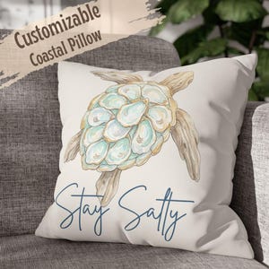 May include: A square off-white throw pillow featuring a central illustration of a sea turtle with a shell composed of oyster shells and driftwood-textured limbs. Below the turtle, the text Stay Salty is printed in a dark blue cursive font.