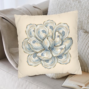 May include: A square throw pillow with a cream-colored background featuring a central illustration of a large, stylized flower composed of overlapping oyster shells in shades of blue, white, and gray, with thin gold-toned outlines on the edges.