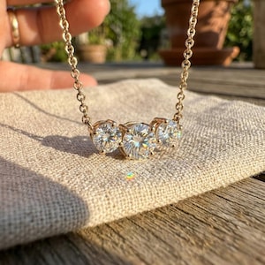 May include: A gold-toned necklace featuring three round, clear gemstones. The necklace is displayed on a beige fabric, with the chain and settings in a matching gold color. The gemstones sparkle, catching the light.