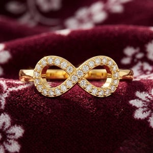 May include: A gold ring featuring an infinity symbol centerpiece encrusted with small, clear, round-cut stones. The band is smooth and polished, connecting to the sides of the infinity design, which is set with multiple sparkling white stones.