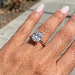 Pode incluir: Um anel de ouro rosa com um grande diamante de corte retangular. O diamante brilha com inúmeras facetas, cravado em uma banda delicada. O anel é mostrado em uma mão com esmalte rosa claro.