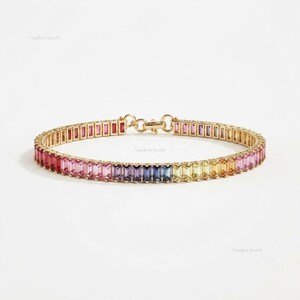 May include: A gold chain bracelet featuring a continuous row of baguette-cut gemstones in a vibrant rainbow gradient. The stones transition through shades of pink, red, purple, blue, yellow, and orange, secured by a gold clasp mechanism.
