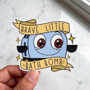 May include: A die-cut sticker featuring a light blue cartoon toaster with large brown eyes and a smiling mouth. The toaster is framed by two yellow banners with the black text BRAVE LITTLE and BATH BOMB, accented by small yellow star icons.