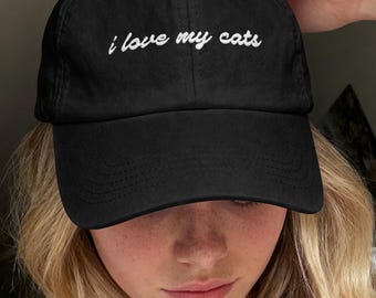 以下が含まれることがあります： フロントパネルに白い刺繍で「i love my cats」と筆記体で書かれた黒いベースボールキャップ。キャップには構造化されたクラウン、カーブしたつば、通気用のステッチ入りアイレットが上部パネルに施されています。