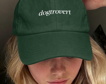 以下が含まれることがあります： 中央に白い小文字のセリフ体で「dogtrovert」という文字が刺繍された、ダークフォレストグリーンの野球帽。この帽子は構造化されたクラウン、カーブしたつば、そしてパネルとつばに沿った目に見えるステッチのディテールが特徴です。
