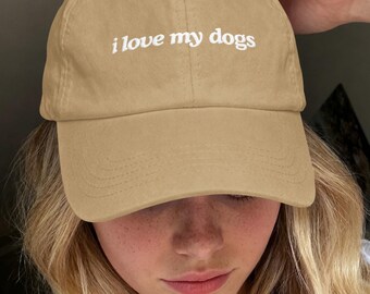 以下が含まれることがあります： フロントパネルの中央に白い刺繍で i love my dogs と書かれたタンカラーの野球帽。カーブしたつば、ステッチ入りのアイレットを備えた構造化されたクラウン、背面に金属製のバックル留め具が付いた布製ストラップが特徴です。