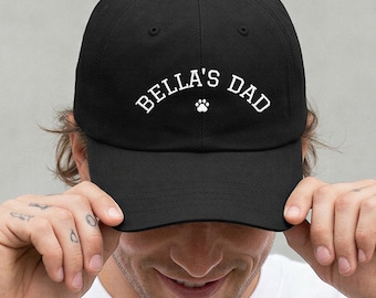 May include: A black baseball cap featuring the white embroidered text BELLA'S DAD in an arched font, centered above a small white paw print icon. The cap has a structured crown, a curved brim, and stitched eyelets on the top panels.