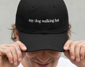 May include: A black baseball cap featuring the white embroidered text "my dog walking hat" centered on the front panel. The cap has a structured crown, a curved brim, and a button at the top, providing a clean and simple design for daily use.