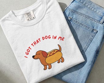 以下が含まれることがあります： 赤い文字で「I GOT THAT DOG IN ME」と書かれた白いTシャツ。ホットドッグのパンに入った漫画のダックスフンドが描かれています。明るい青色のデニムジーンズが隣にあります。