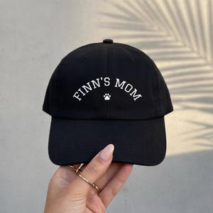 A black baseball cap featuring the white embroidered text FINN'S MOM in an arched font, centered above a small white paw print icon. The cap has a structured crown, a curved brim, and a button at the top center of the fabric panels.