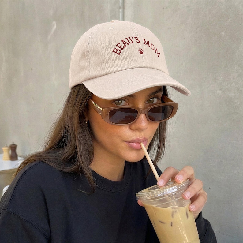 A beige baseball cap featuring the embroidered text BEAU'S MOM in dark red lettering with a small paw print icon centered below the text. The cap has a structured crown, a curved brim, and a fabric strap closure at the back.