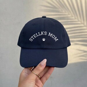 A navy blue baseball cap featuring the white embroidered text STELLA'S MOM in an arched layout, centered above a small white paw print icon. The cap has a structured crown, a curved brim, and a button at the top center of the crown.