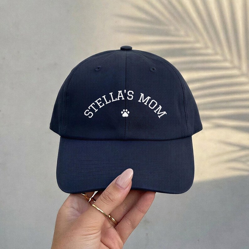 A navy blue baseball cap featuring the white embroidered text STELLA'S MOM in an arched layout, centered above a small white paw print icon. The cap has a structured crown, a curved brim, and a button at the top center of the crown.