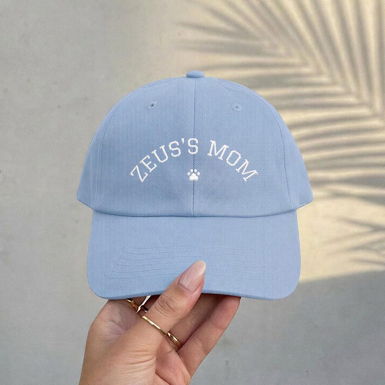 A light blue baseball cap featuring the white embroidered text ZEUS'S MOM in an arched layout, centered above a small white paw print icon. The cap has a structured crown, a curved brim, and visible stitching details on the front panels.
