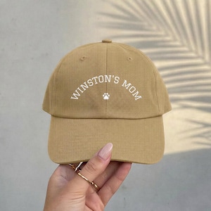 A tan baseball cap featuring the white embroidered text WINSTON'S MOM in an arched design, centered above a small white paw print icon. The cap has a structured crown, a curved brim, and visible stitching details across the fabric panels.