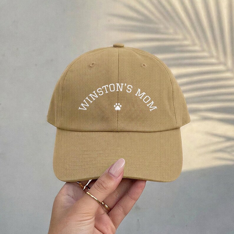 A tan baseball cap featuring the white embroidered text WINSTON'S MOM in an arched design, centered above a small white paw print icon. The cap has a structured crown, a curved brim, and visible stitching details across the fabric panels.