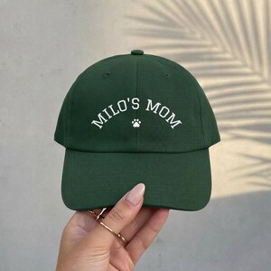 A dark forest green baseball cap featuring the white embroidered text "MILO'S MOM" in an arched font, centered above a small white paw print icon. The cap has a structured crown, a curved brim, and a button at the top center.