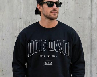 以下が含まれることがあります： 大胆なカレッジフォントでDOG DADと白くプリントされた黒のクルーネックトレーナー。テキストの下には、中央に小さな肉球のアイコンを添えてESTD 2024と記され、その下に枠で囲まれたBAILEYという名前が配置されています。