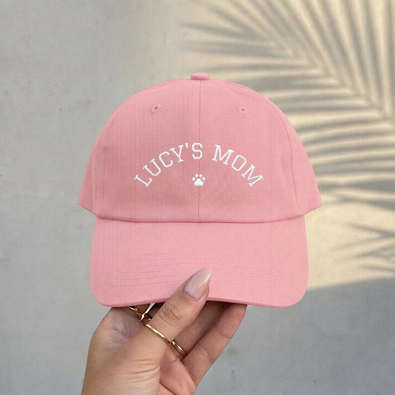 A light pink baseball cap featuring the white embroidered text "LUCY'S MOM" in an arched font, centered above a small white paw print icon. The cap has a structured crown, a curved brim, and a button at the top center of the fabric.