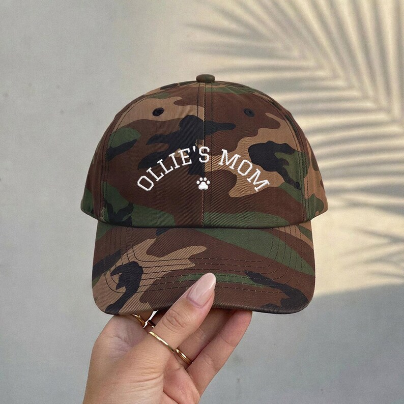 A camouflage baseball cap featuring a brown, green, and black pattern. The front of the cap displays the white text OLLIE'S MOM in an arched font, positioned above a small white paw print icon centered on the crown.