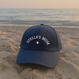 A navy blue baseball cap featuring white embroidered text that reads STELLA'S MOM with a small white paw print icon centered below the text. The cap has a structured crown, a curved brim, and stitched eyelets on the top panels.