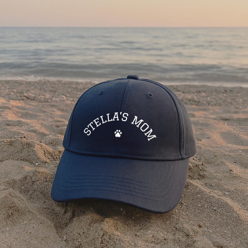 A navy blue baseball cap featuring white embroidered text that reads STELLA'S MOM with a small white paw print icon centered below the text. The cap has a structured crown, a curved brim, and stitched eyelets on the top panels.