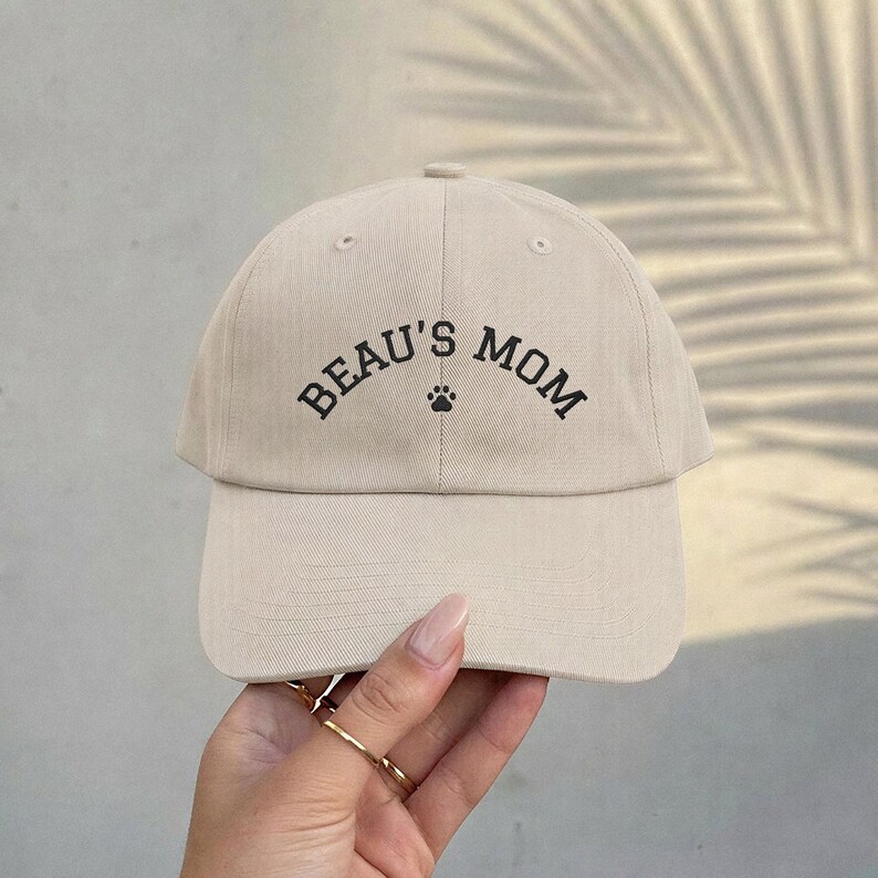 A beige baseball cap featuring the black embroidered text BEAU'S MOM in an arched font, with a small black paw print icon centered between the words. The cap has a structured crown, a curved brim, and a button on the top center.