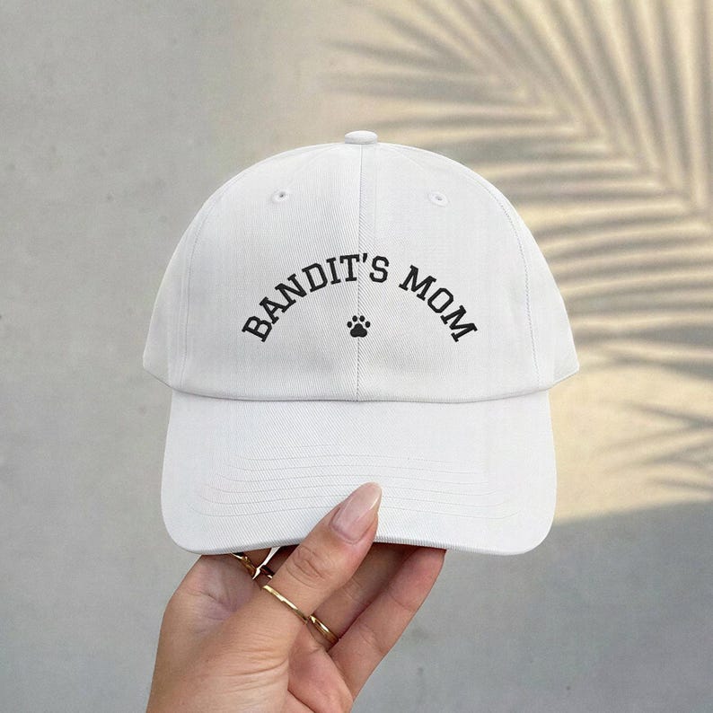 A white baseball cap featuring the black text BANDIT'S MOM arched over a small black paw print icon. The cap has a structured crown, a curved brim, and a button at the top center, constructed from a solid white fabric material.