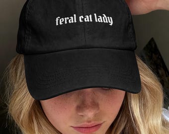 May include: A black baseball cap featuring the text "feral cat lady" embroidered in white, gothic-style lettering across the front panel. The cap has a curved brim and a structured crown with visible stitching details on the fabric surface.