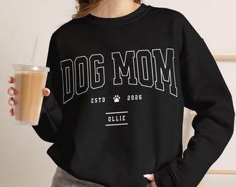 以下が含まれることがあります： DOG MOMと書かれた白いカレッジスタイルの文字が特徴の黒いクルーネックのスウェットシャツ。テキストの下には、ESTDという単語、小さな肉球のアイコン、2026年という年号、そして2本の水平線で下線が引かれたOLLIEという名前がデザインされています。