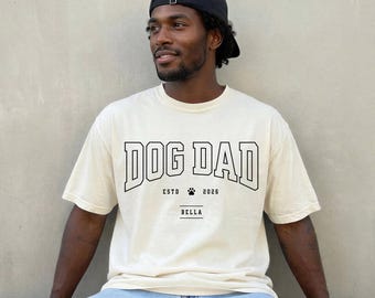 以下が含まれることがあります： オフホワイトの半袖Tシャツ。カレッジフォントで黒い文字の「DOG DAD」がプリントされ、その下に「ESTD 2026」、小さな肉球のアイコン、そして小さめのフォントで「BELLA」という名前が中央に配置されています。黒いベースボールキャップを着用しています。