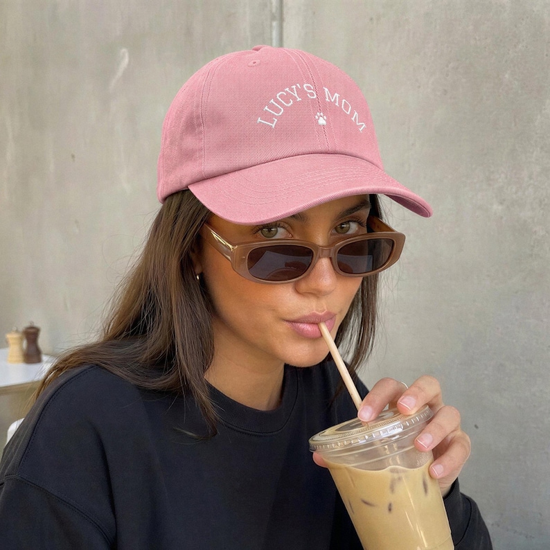 A dusty pink baseball cap featuring the white embroidered text LUCY'S MOM with a small paw print icon centered below the text. The cap has a structured crown and a curved brim, constructed from a solid fabric material.