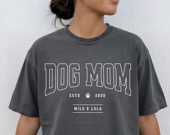 以下が含まれることがあります： チャコールグレーの半袖Tシャツ。大学風のフォントで白く「DOG MOM」とプリントされています。その下には「ESTD 2026」と肉球のアイコンがあり、さらにその下に2本の水平線の間に中央揃えで「MILO & LOLA」という名前が記されています。