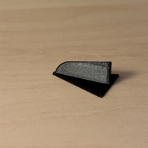 K&ouml;nnte beinhalten: Ein kleines, schwarzes, dreieckiges Objekt mit einem strukturierten, grauen, rechteckigen Teil. Das Objekt liegt auf einer hellbraunen Oberfl&auml;che. Das Objekt scheint 3D-gedruckt zu sein.